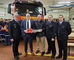 Fünf Männer, drei davon in Feuerwehruniform stehen vor einem roten Feuerwehrauto und halten einen großen roten Schlüssel in die Hand. Im Hintergrund stehen Mitglieder der Jugendfeuerwehr in neonfarbenen Jacken.